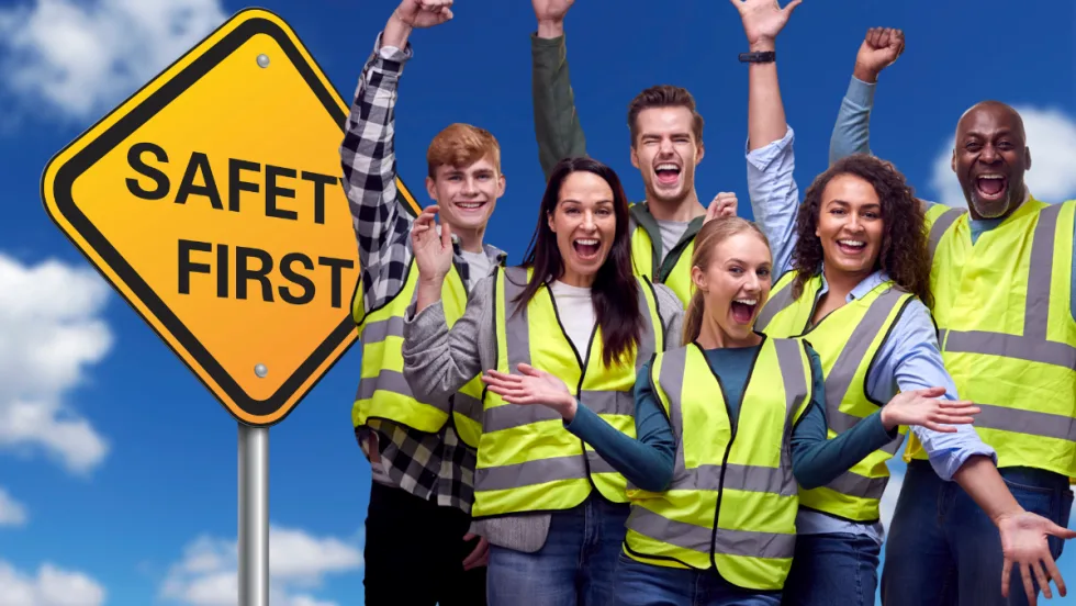 Group of smiling workers in safety vests standing by a Safety First sign, celebrating a strong workplace safety culture.