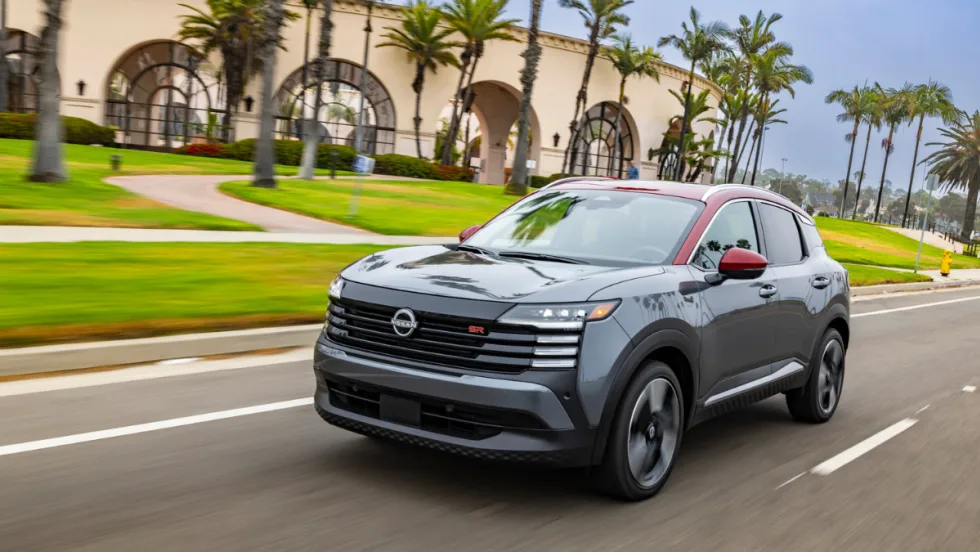 A 2026 Nissan Kicks SR in gray with red accents drives on a palm-lined street, showcasing the SUV’s updated styling and compact crossover design.