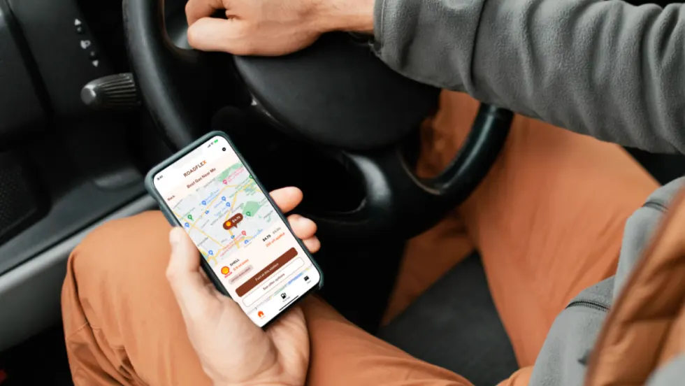 Driver holding a phone with the RoadFlex fuel management app open, showing a map with gas stations and real-time prices inside a vehicle.