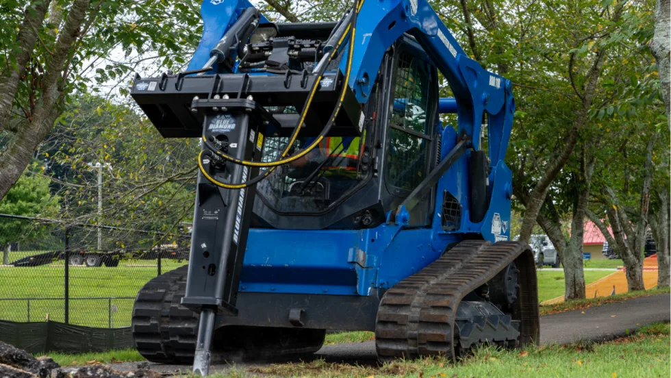 Blue skid steer using a hydraulic breaker to demolish paved sidewalk in a park setting.