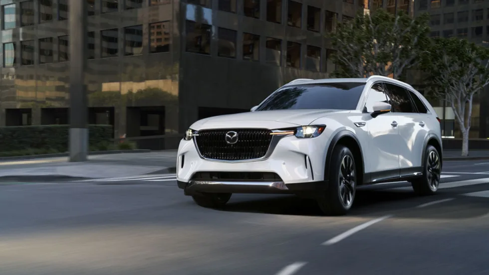 A white 2026 Mazda CX-90 crossover SUV driving through a city street, showcasing its front grille, LED headlights, and modern exterior design.