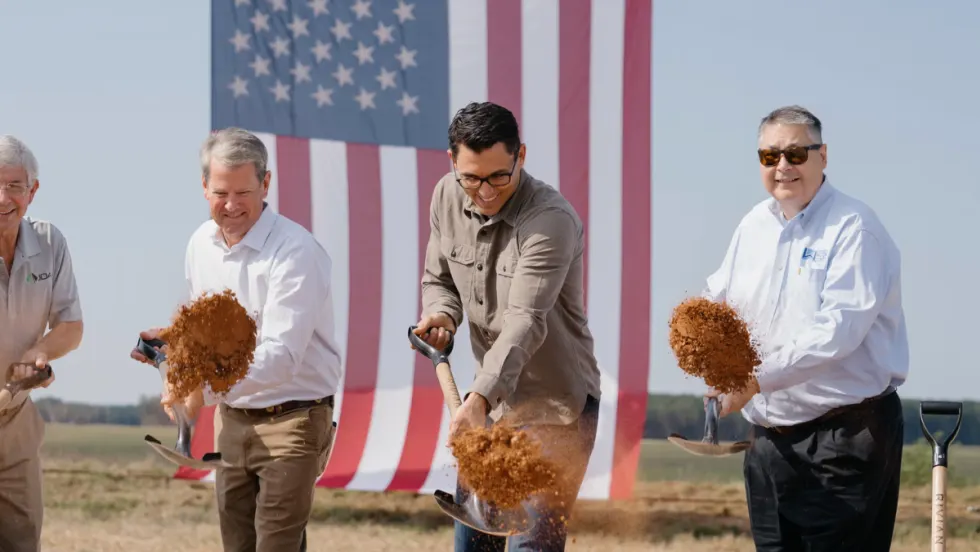 Rivian CEO RJ Scaringe, Georgia Governor Brian Kemp, and other officials shovel dirt during the groundbreaking ceremony, with a large American flag in the background.