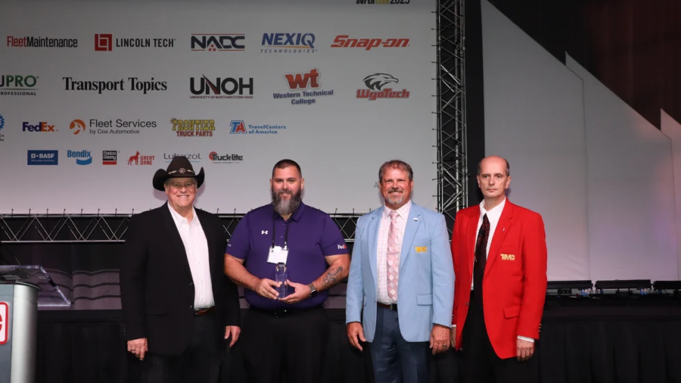Matthew Sloan, winner of TMCSuperTech 2025, stands with event officials and holds his award during the recognition ceremony, with sponsor logos displayed on the backdrop.
