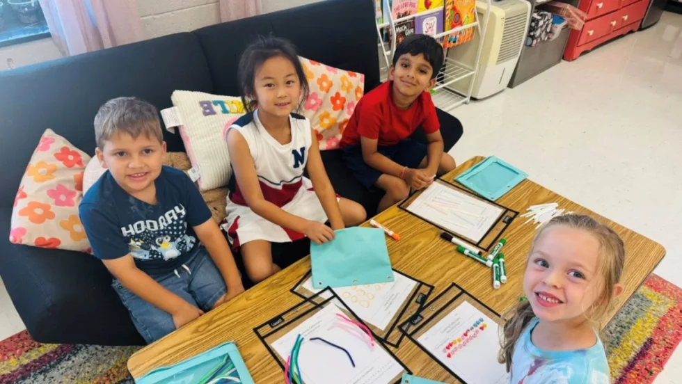 Four young students sit at a classroom table with worksheets, markers, and colorful pipe cleaners, smiling while engaging in a STEM activity.