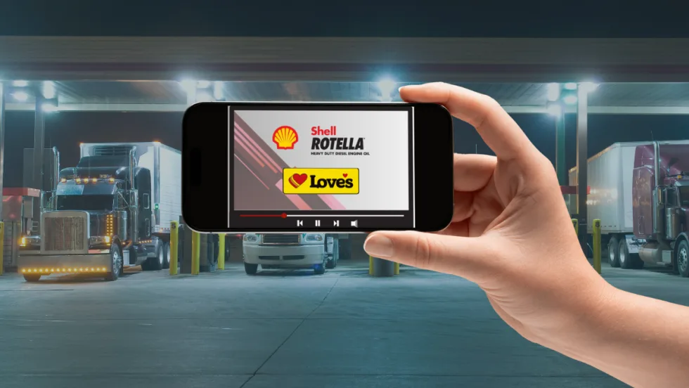 Hand holding a smartphone displaying a Shell Rotella and Love’s Travel Stops video screen, with semi-trucks parked at a brightly lit fuel station in the background.