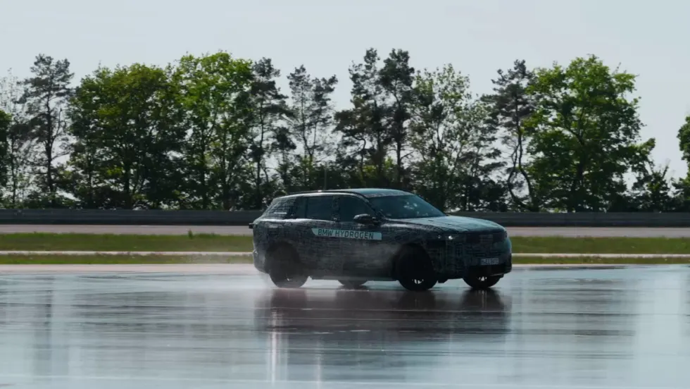 Camouflaged BMW iX5 Hydrogen SUV driving on a wet test track, kicking up water spray, with trees and a racetrack barrier in the background.