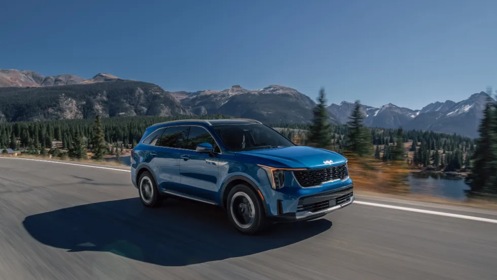 Blue 2026 Kia Sorento Hybrid driving on a mountain highway with trees and water in the background.