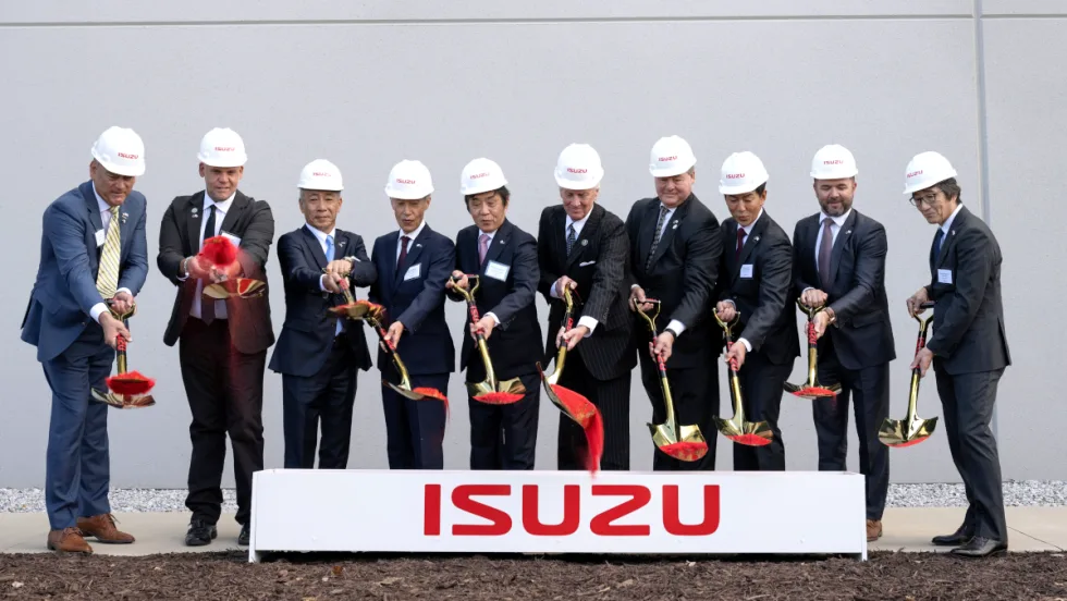 Business men stand behind a box with Isuzu logo and shovel dirt onto the ground.
