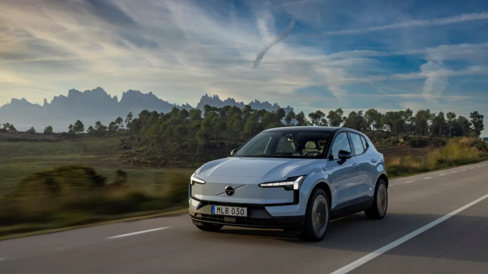 A light blue 2026 Volvo EX30 electric SUV driving on a rural highway with mountains and trees in the background under a clear sky.
