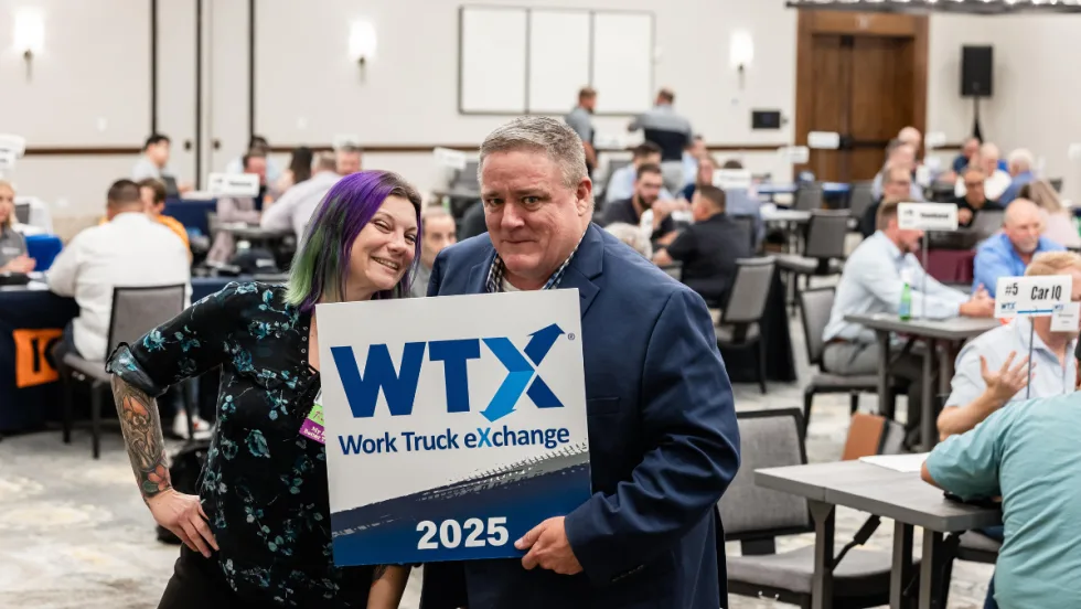 Lauren Fletcher and Wayne Parham posing with an attendee holding a Work Truck Exchange 2025 sign inside the meeting room filled with one-on-one business meetings.