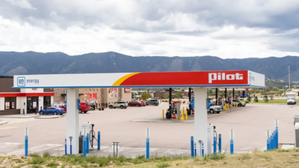 A Pilot Travel Center with GM Energy and EVgo fast-charging stations installed under the canopy, with fuel pumps and vehicles in the background against a mountain landscape.