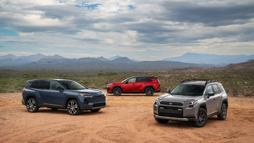 Three 2026 Toyota RAV4 SUVs—gray, red, and blue—parked in a desert landscape with mountains in the distance.