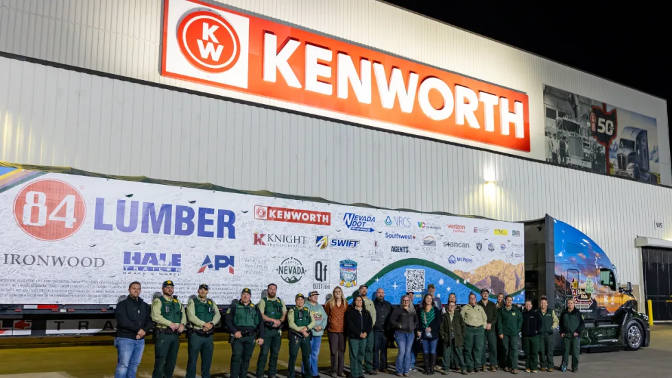 Group of people stand in front of a tractor trailer parked outside a building with large Kenworth sign