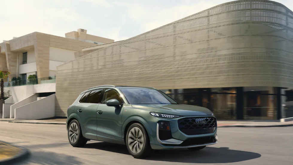 Green 2026 Audi Q3 compact SUV driving on a city street past a modern building in daylight.