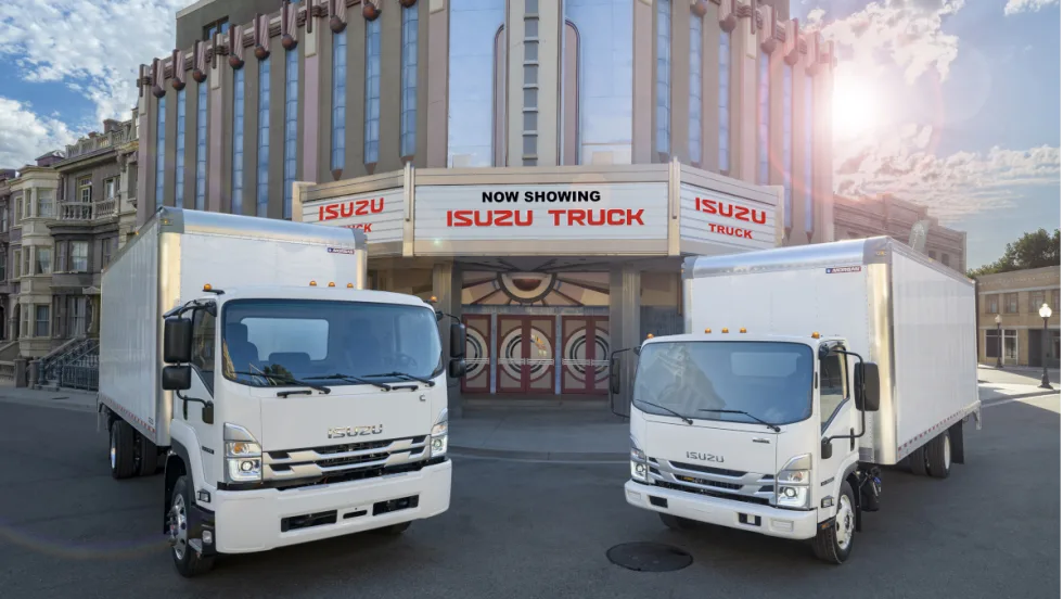 Two Isuzu box trucks parked in front of a movie theater with sign that says Now Showing, Isuzu Truck.