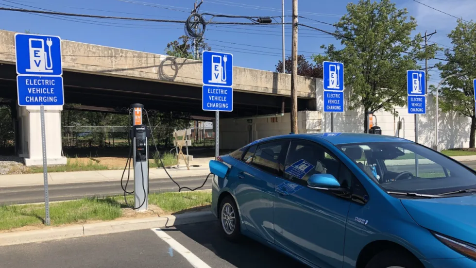 Blue electric car plugged into an EV charging station beneath an overpass, with multiple “Electric Vehicle Charging” parking signs.