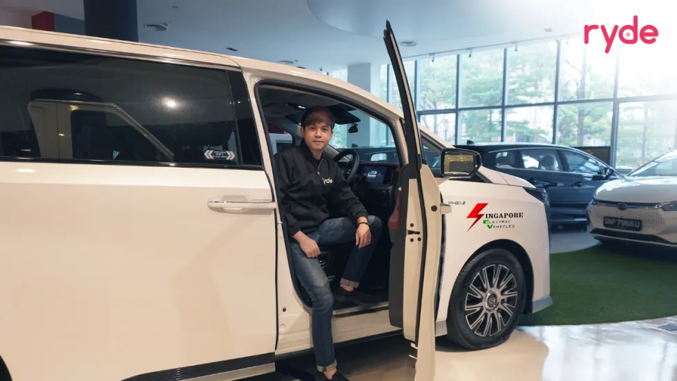 Ryde driver seated in a white electric vehicle inside a showroom, with Singapore Electric Vehicles branding on the car and other vehicles in the background.