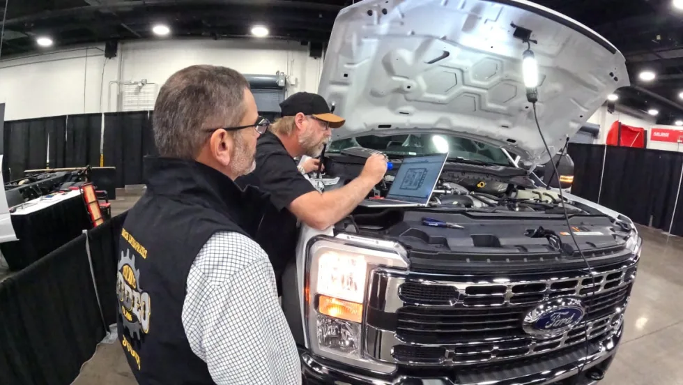 One man watching another work under the hood of a large pickup truck.