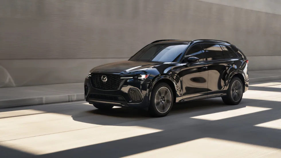 A black Mazda CX-70 crossover driving through an urban roadway with concrete walls in the background.