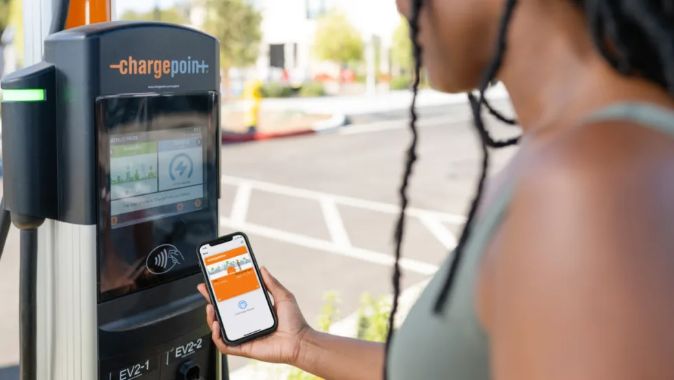 A driver uses the ChargePoint app on a smartphone next to a ChargePoint EV charging station.