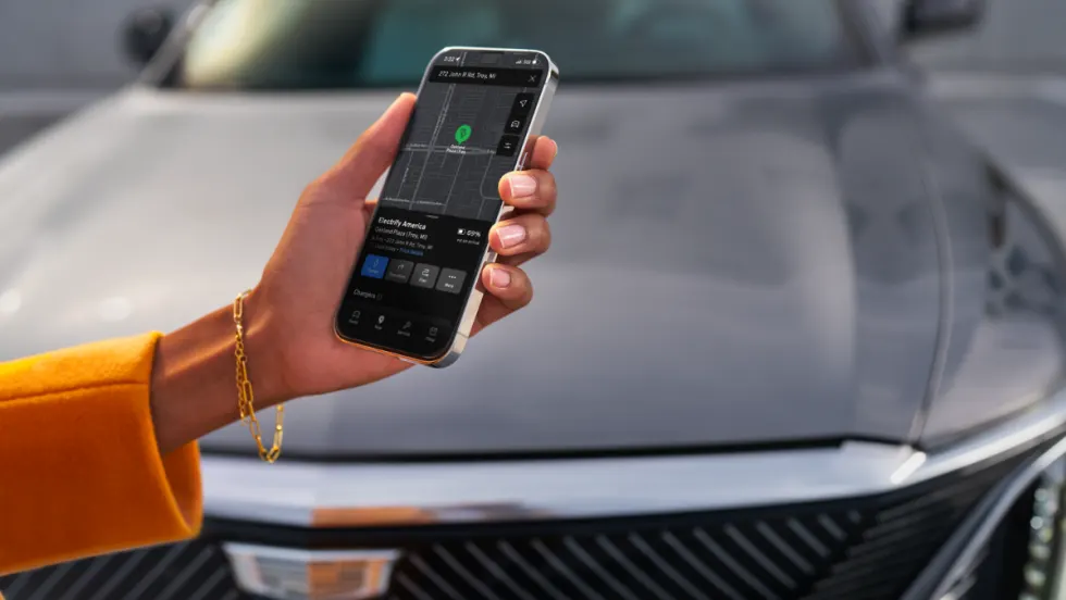 A driver holds a smartphone displaying a GM vehicle app with an Electrify America charging station map, with an electric vehicle parked in the background.