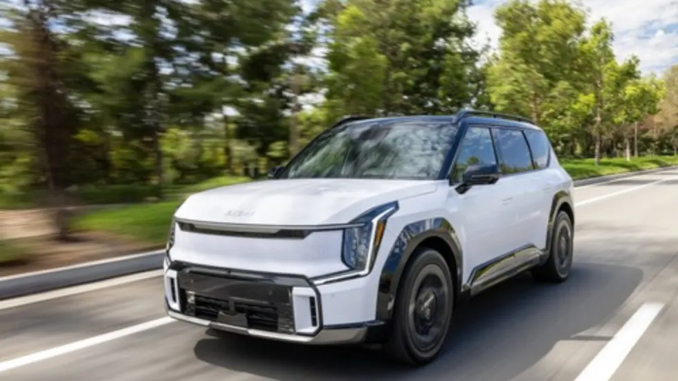 A white 2026 Kia EV9 electric SUV driving on a tree-lined roadway.
