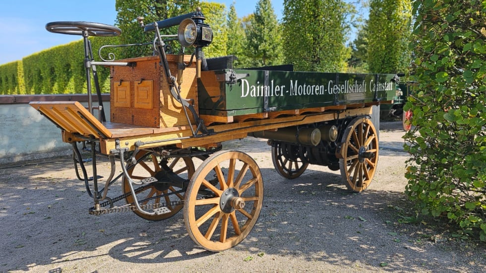 Reconstructed 1896 Daimler motorized truck with wooden wheels and rear-mounted engine displayed outdoors.