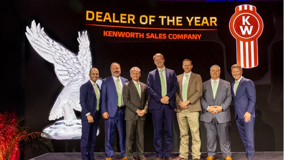 Group of men in business attire on a stage during an award ceremony with background that says Dealer of the Year and a Kenworth logo.