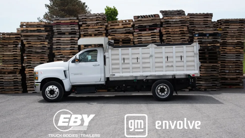 Side view photo of a white stake-bed work truck with logos across the bottom of the image for EBY and GM Envolve.