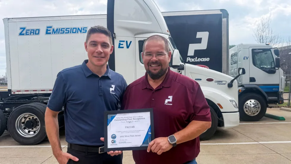Two men stand together holding a certificate award with large commercial trucks in the background.