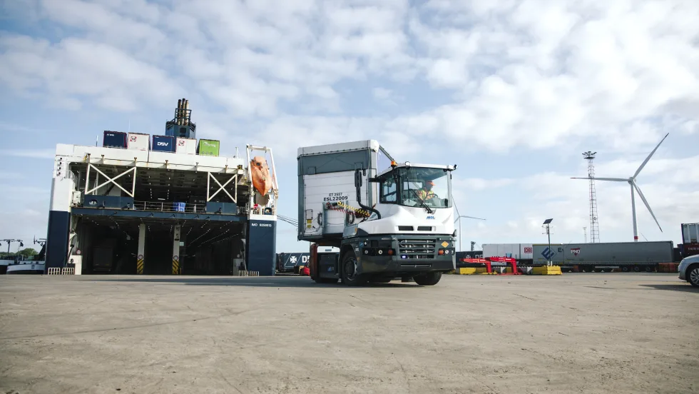 Terminal truck hauling a container trailer with a cargo ship and windmill in the background.