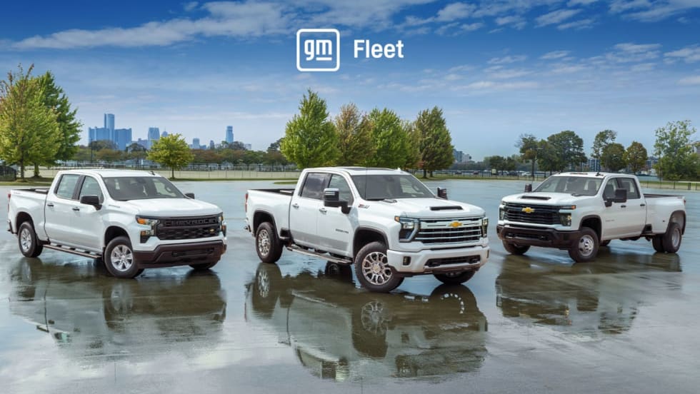 Three white pickup trucks parked in a parking lot with trees in background and a blue sky with logo for GM Fleet.