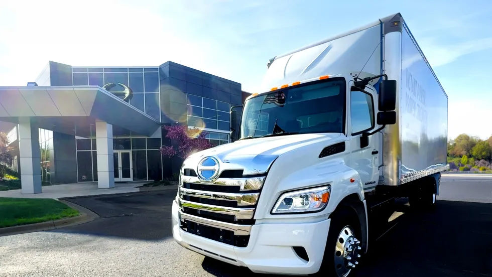 White box delivery truck parked outside a modern office building on a sunny day. The truck is photographed from the front angle, with chrome detailing reflecting sunlight and the building entrance visible in the background.