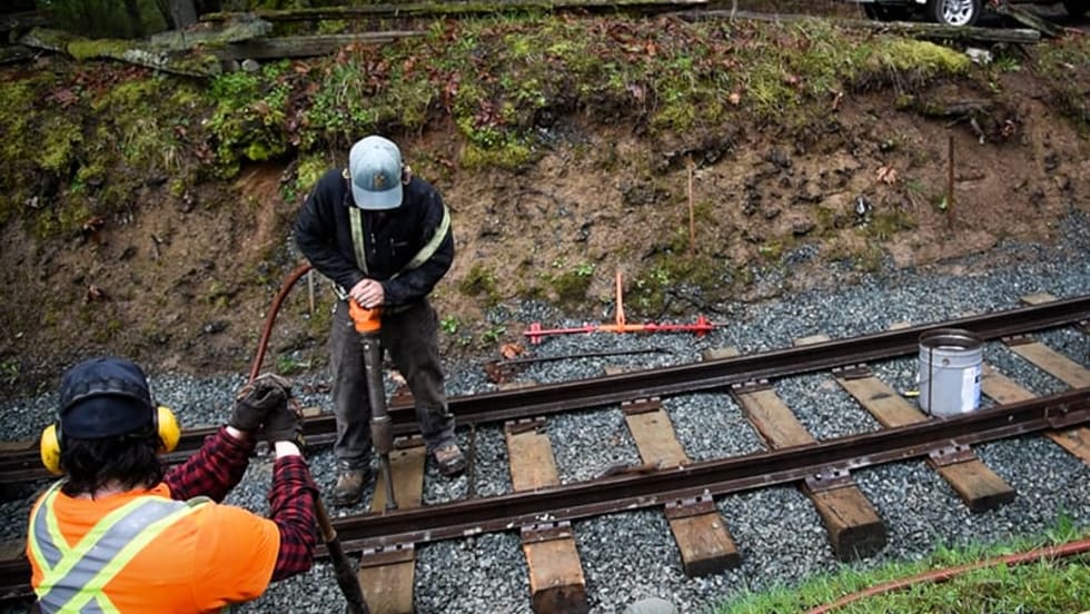 Two workers in safety gear perform maintenance on railroad tracks in a wooded outdoor area. One operates heavy equipment on the rails while another observes nearby, with a white utility truck parked above the embankment.