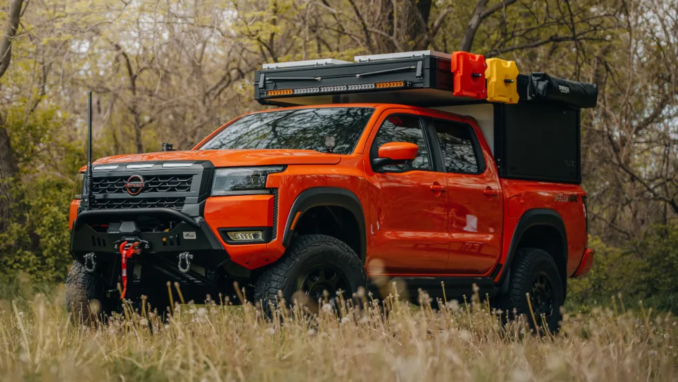 Orange Nissan Frontier PRO-4X overland build parked in a grassy wooded area with off-road tires, heavy-duty front bumper, winch, rooftop cargo storage, and camping accessories mounted above the truck bed.