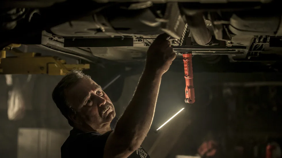 Mechanic working beneath a raised vehicle in a dimly lit garage, illuminated by a hanging Streamlight work light. The technician reaches toward the undercarriage while inspecting or repairing automotive components.
