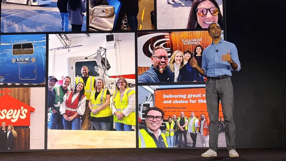 Presenter speaking onstage in front of a large display wall featuring fleet and transportation team photos, including trucks, drivers, and company groups wearing safety vests.