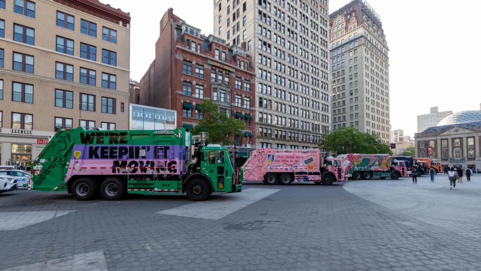 Line of brightly painted New York City sanitation trucks displayed in a public plaza, featuring colorful murals and messages. Tall city buildings and pedestrians surround the outdoor exhibition in an urban setting.