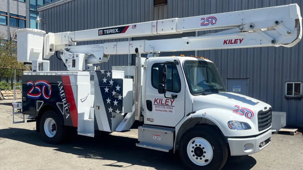 White Kiley utility truck equipped with a Terex aerial lift parked outside an industrial building. The truck features patriotic red, white, and blue graphics, including an American flag design and “250” anniversary emblems on the body and boom.
