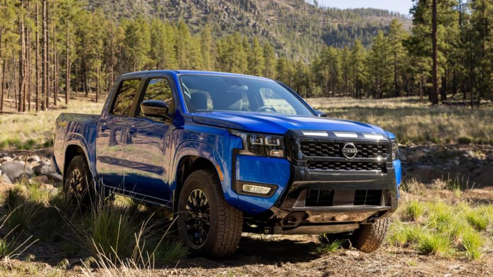 Blue Nissan Frontier pickup truck parked on rugged terrain in a forested mountain landscape. The four-door truck is shown from the front three-quarter angle, highlighting its off-road stance, black grille, and outdoor adventure setting.