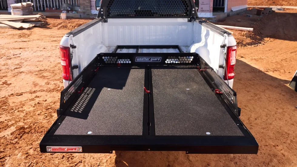 Rear view of a white work truck at a construction site featuring a Weather Guard sliding truck-bed system extended outward, providing easy access to tools and equipment. A protective cab rack and unfinished building frame the background.