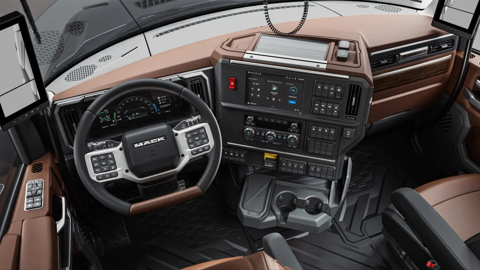 Driver’s-eye view of a Mack Granite truck interior featuring a digital instrument cluster, multifunction steering wheel, large touchscreen infotainment system, integrated controls, cup holders, and premium brown-and-black trim designed for comfort and productivity.