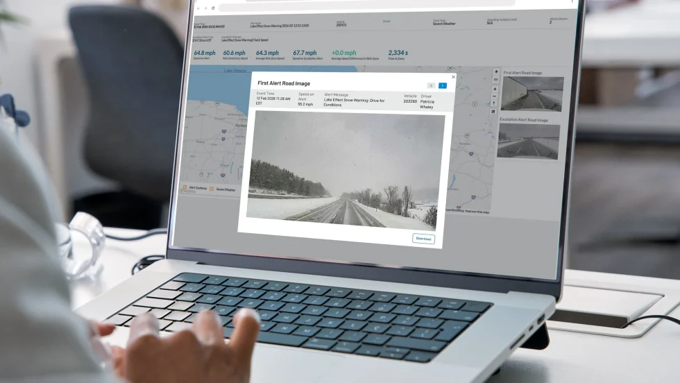Person using a laptop to view a fleet management dashboard displaying weather alerts, vehicle speed data, maps, and road-condition images. The screen highlights a snowy roadway and a lake-effect snow warning for a fleet vehicle.