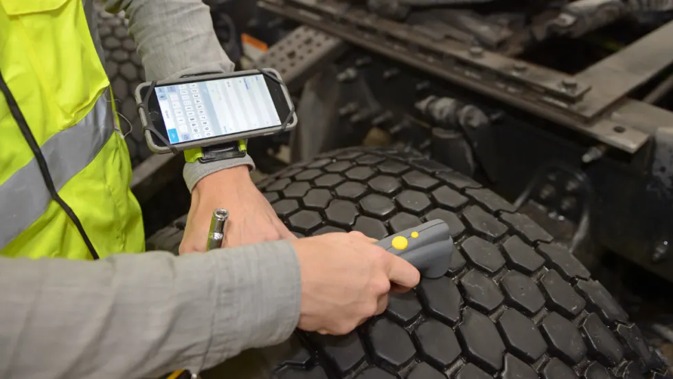 Technician wearing a high-visibility safety vest uses a handheld scanning device to inspect a commercial truck tire while viewing data on a wrist-mounted tablet, illustrating digital tire maintenance and wheel torque tracking technology.
