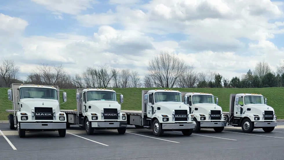 Five white Mack MD-series flatbed trucks are parked side by side in a lot under a cloudy sky, showcasing a fleet of medium-duty commercial vehicles with aluminum flatbed bodies.