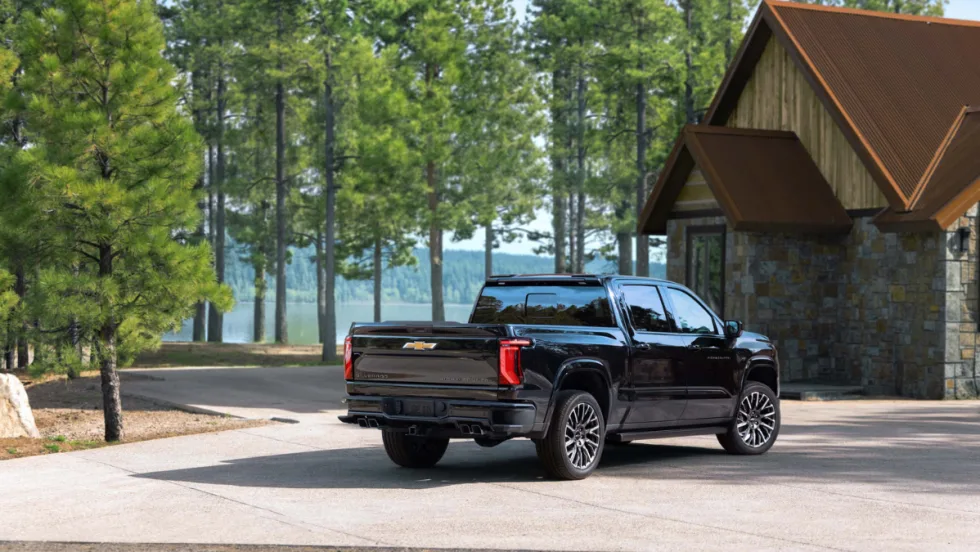 Rear three-quarter view of a black 2027 Chevrolet Silverado 1500 parked outside a lakeside cabin surrounded by pine trees.