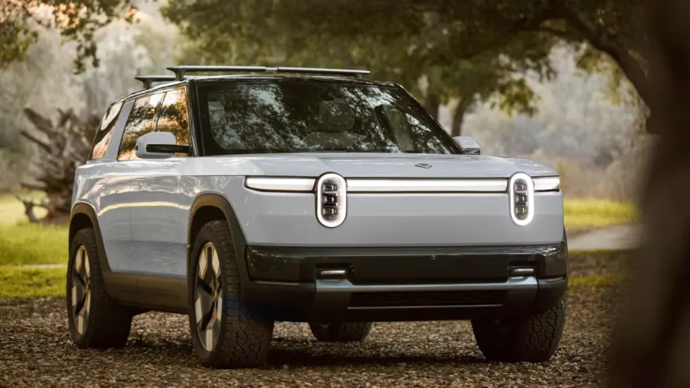 White Rivian R2 electric SUV parked on a leaf-covered path in a wooded area, shown from the front three-quarter angle with distinctive oval headlights, black trim, and roof rails under soft natural light.