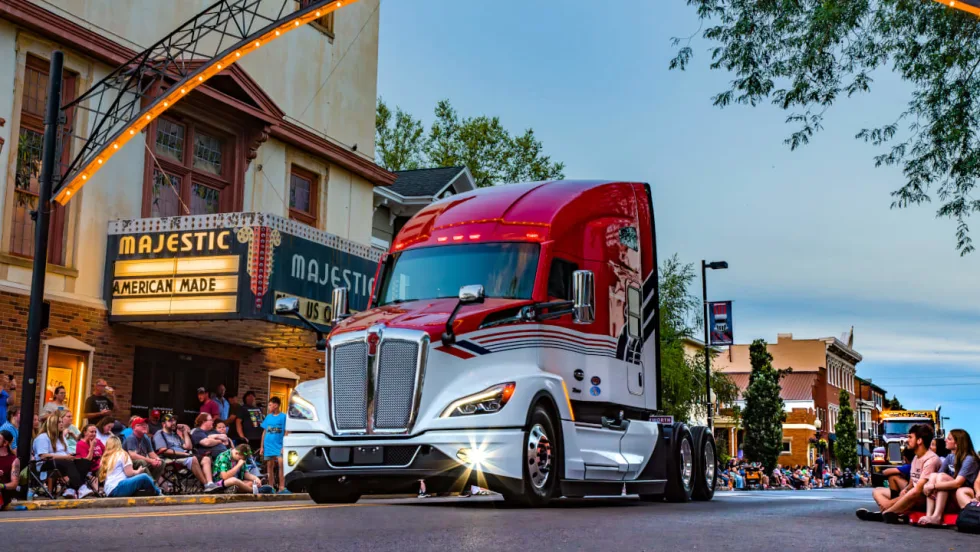 Red-and-white Kenworth semi-truck drives through a small-town parade at dusk. Spectators line the street outside the Majestic Theater as another decorated truck follows in the background.