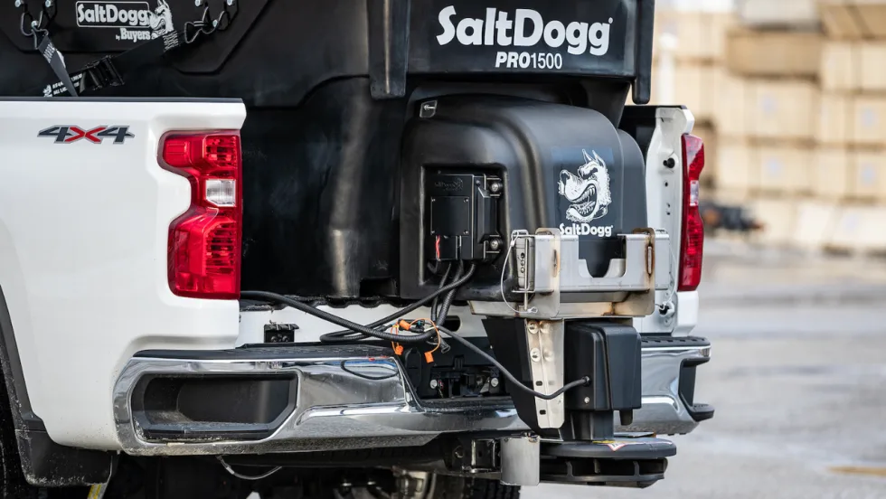 Close-up of a SaltDogg PRO1500 tailgate salt spreader mounted on the rear of a white 4x4 pickup truck, showing the hopper, spreader assembly, wiring connections, and mounting hardware.