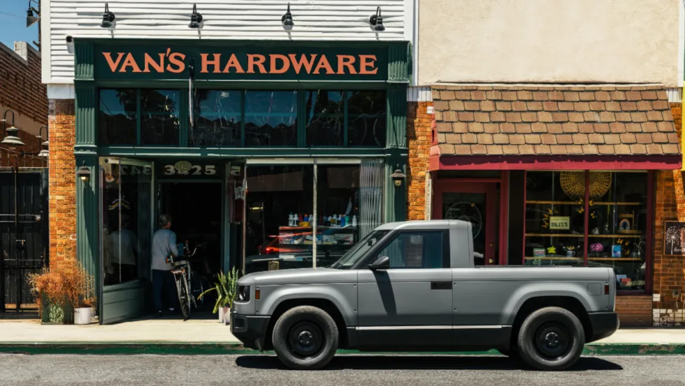 Gray Slate electric pickup truck parked curbside in front of Van’s Hardware on a small-town main street, highlighting the truck’s minimalist design and compact single-cab configuration.